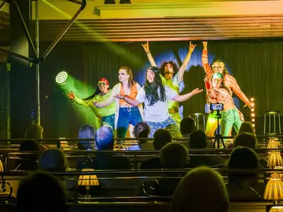 Eine Gruppe von fünf jungen Darstellern in farbenfrohen Hippie-Kostümen performt auf einer Bühne an Bord der MS Deutschland. Das Publikum ist im Vordergrund sitzend erkennbar.