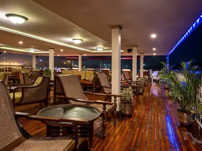 Die überdachte Bar-Lounge der MS Ganges Voyager bei Nacht. Elegante Rattanmöbel, warmes Deckenlicht und blaue LED-Beleuchtung schaffen ein gemütliches Ambiente. Topfpflanzen und eine beleuchtete Küstenlinie sind sichtbar.