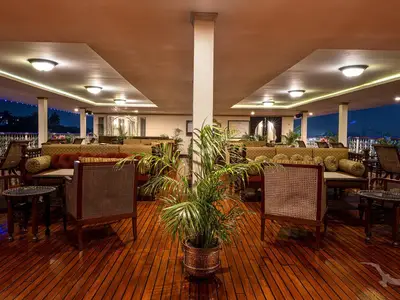 Eine elegante Bar-Lounge an Bord der MS Ganges Voyager am Abend. Gemütliche Sofas und Sessel mit Rattangeflecht sind auf dunklem Holzdeck arrangiert. Im Hintergrund leuchten Lichter am Ufer und im Inneren des Schiffs. Eine Palme steht zentral.