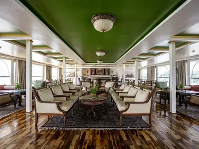 Elegante Bar-Lounge der MS Ganges Voyager. Gemütliche Sitzgruppen mit hellen Sesseln und Holztischen. Im Hintergrund die Bar. Große Fenster mit weitem Ausblick. Grüne Decke, Holzboden.