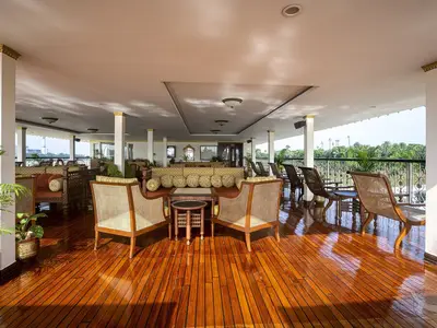Geräumige, offene Lounge an Bord der MS Ganges Voyager mit glänzendem Holzboden, eleganten Rattanmöbeln und Sofas. Panoramablick auf Fluss und üppige Vegetation.