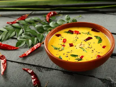 Nahaufnahme einer cremigen, gelben Kokossuppe in einer braunen Schale auf einem dunklen Holztisch, garniert mit roten Chilis, Curryblättern und Senfsaat. Im Hintergrund weitere Chilis und Curryzweige. Gesehen an Bord der MS Ganges Voyager.