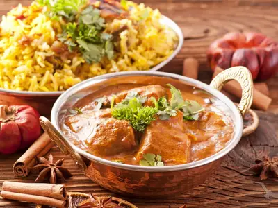 Indisches Hühnchen-Curry in Kupferschale, garniert mit frischem Koriander. Dazu gelber Reis und Gewürze wie Zimt und Sternanis auf Holztisch. Serviert an Bord der MS Ganges Voyager.