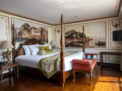 Eine elegant eingerichtete Suite an Bord der MS Ganges Voyager mit einem Himmelbett, reich verzierten Möbeln und Wandgemälden, die Flusslandschaften zeigen. Ein kleiner Sessel steht im Vordergrund.