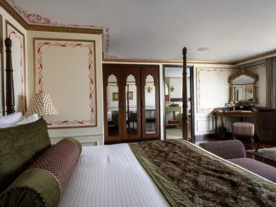 Detailreiche Suite an Bord der MS Ganges Voyager. Himmelbett mit grünen Kissen, kunstvolle rote Wandornamente und ein Holztisch mit großem goldenem Spiegel. Klassisch-luxuriöses Interieur.