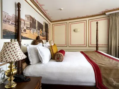 Die luxuriöse Suite der MS Ganges Voyager zeigt ein Himmelbett mit weißer Bettwäsche und rot-goldenen Zierkissen. Opulent dekorierte Wände mit einem großen Gemälde und roten Bordüren umrahmen den Raum. Eine Messinglampe beleuchtet den Nachttisch.