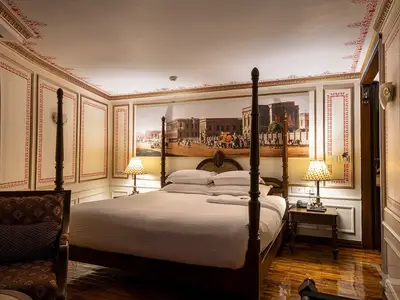 Eine elegante Kabine an Bord der MS Ganges Voyager. Im Zentrum ein komfortables Himmelbett mit weißen Laken und Kissen, flankiert von zwei Nachttischen mit Tischlampen. Über dem Bett hängt ein breites Wandgemälde mit historischen Gebäuden und Menschen. Die Wände sind mit roten Ornamenten verziert.