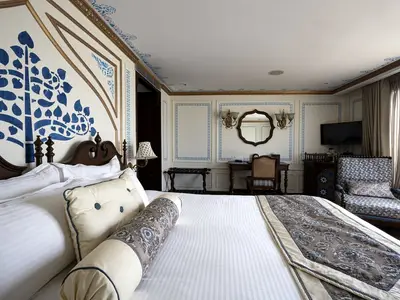 Helle, elegante Suite an Bord der MS Ganges Voyager. Ein großes Bett mit weißer Bettwäsche steht links. Rechts sind ein Schreibtisch, Spiegel und ein Sessel. Die Wände sind in Weiß und Blau verziert.
