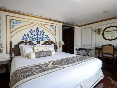 Elegante Suite an Bord der MS Ganges Voyager. Im Zentrum ein Doppelbett mit kunstvollem Kopfteil, umliegende Wände in Blau und Gold verziert. Rechts ein Schreibtisch mit Stuhl und Spiegel.