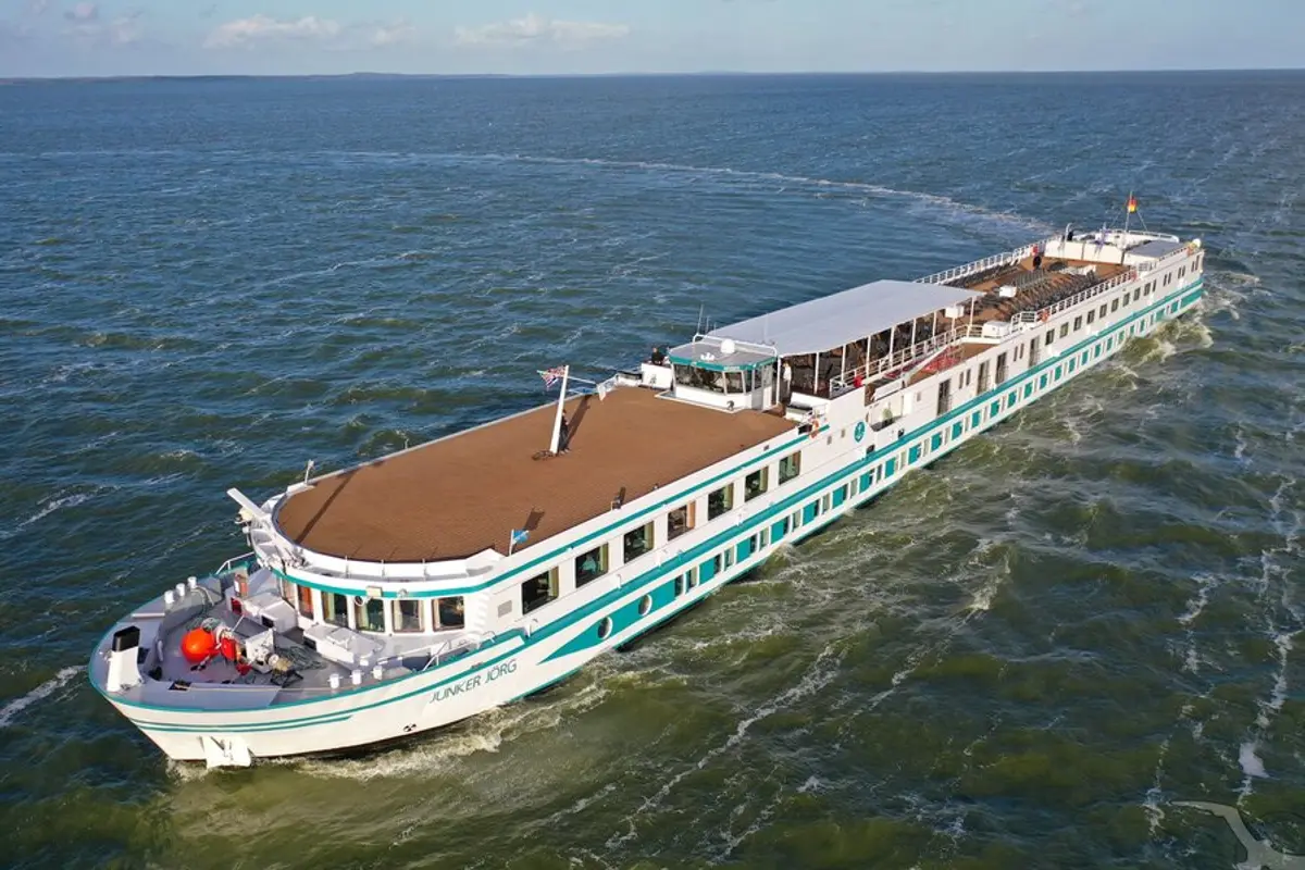 Die MS Junker Jörg, ein weißes Flusskreuzfahrtschiff mit türkisgrünen Streifen, fährt auf leicht welligem, blauem Wasser. Klare Außenansicht, von oben aufgenommen bei hellem Tageslicht.