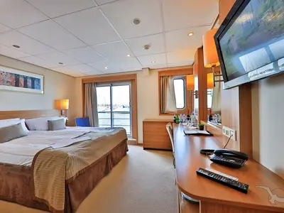 Innenansicht einer geräumigen Balkonkabine an Bord der MS Junker Jörg mit Doppelbett, Schreibtisch und Ausblick auf den Hafen durch eine Glasschiebetür und ein Seitenfenster.
