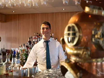 Ein freundlicher Barkeeper lächelt hinter einer gut bestückten Bar an Bord der MS Junker Jörg. Im Vordergrund rechts ist unscharf ein kupferner Taucherhelm zu sehen.