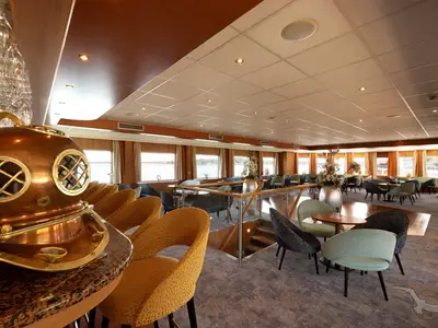 Die Bar-Lounge an Bord der MS Junker Jörg mit verschiedenen Sitzgruppen, großen Fenstern und einem dekorativen Taucherhelm auf der Theke. Einladende Atmosphäre mit Blick auf das Wasser.