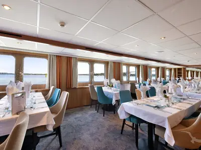 Das helle, moderne Restaurant der MS Junker Jörg zeigt elegant gedeckte Tische mit weißen Tischdecken und teal Stühlen. Große Fenster bieten einen malerischen Blick auf eine ruhige Wasserlandschaft mit grünen Ufern und einem blauen Himmel.