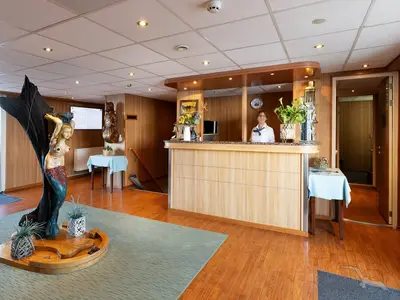 Die Rezeption der MS Junker Jörg mit einer lächelnden Empfangsdame. Der Bereich ist mit hellem Holz verkleidet und nautischen Elementen, darunter eine Meerjungfrauen-Skulptur und ein Leuchtturmmodell, dekoriert.