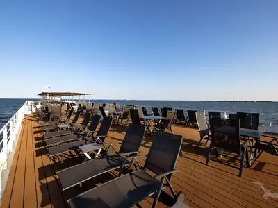 Ein weitläufiges Sonnendeck der MS Junker Jörg mit vielen schwarzen Liegestühlen und Esstischen unter blauem Himmel und Blick auf das weite Meer. Keine Personen sichtbar.