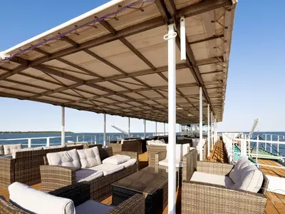 Überdachtes Sonnendeck der MS Junker Jörg mit Rattan-Loungegruppen und hellen Polstern. Im Hintergrund: Meer, Küste und blauer Himmel.