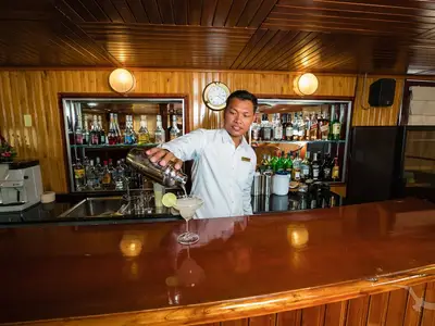 Ein Barkeeper der MS Lan Diep gießt einen Cocktail in ein Glas. Die Bar ist holzvertäfelt, mit Regalen voller Spirituosen und einer glänzenden Theke. Eine einladende Szene.