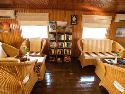 Die helle, holzvertäfelte Lounge der MS Lan Diep zeigt Rattanmöbel mit gestreiften Kissen, Glastische und ein Bücherregal, alles beleuchtet durch große Fenster.