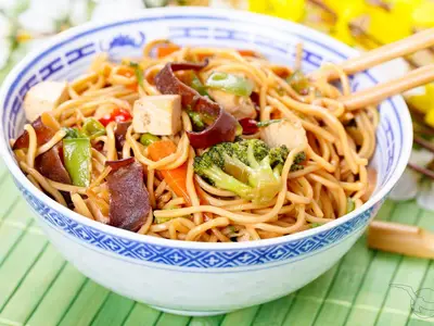 Nahaufnahme eines asiatischen Nudelgerichts mit Tofu, Brokkoli, Karotten und Pilzen in einer blau-weiß gemusterten Schale. Essstäbchen sind sichtbar. Serviert an Bord der MS Lan Diep.