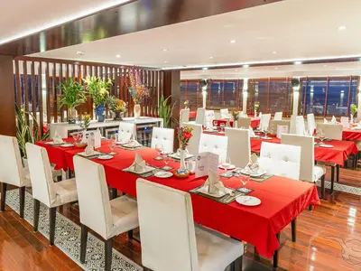 Elegantes Restaurant an Bord der MS Lan Diep. Mit roten Tischdecken gedeckte Tische, weiße Stühle und frische Blumendeko. Große Fenster mit Holzjalousien im Hintergrund.