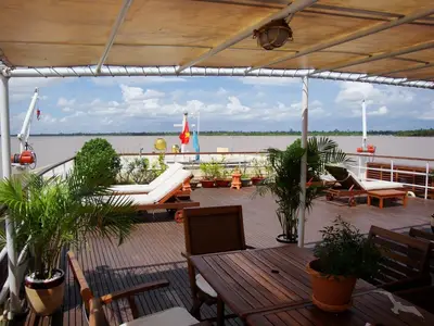 Das Sonnendeck der MS Lan Diep zeigt Holzmöbel, Pflanzen und Liegestühle unter einem Sonnensegel. Ein breiter Fluss und blauer Himmel im Hintergrund.