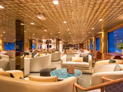 Eine großzügige Lounge an Bord der MS Magellan in warmer Abendbeleuchtung. Bequeme helle Sofas und Sessel laden zum Verweilen ein. Große Fenster geben den Blick nach draußen frei. Die Decke ist mit vielen Lichtern versehen.