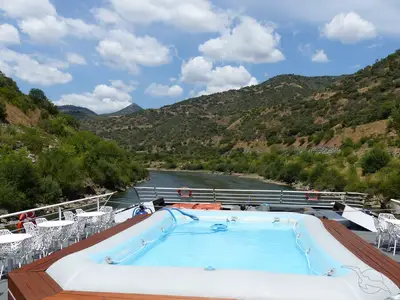 Sonnendeck der MS Magellan mit blauem Pool und weißen Tischen. Im Hintergrund fliesst ein Fluss durch bewaldete Hügel unter blauem Himmel.