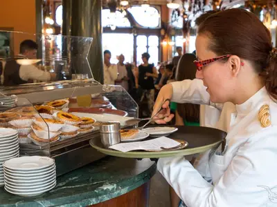 Eine Servicekraft der MS Magellan mit roter Brille serviert Pasteis de Nata von einer Auslage auf ein Tablett. Im Hintergrund sind weitere Gäste und ein Mitarbeiter zu sehen.