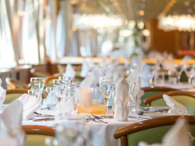Ein festlich gedeckter Tisch mit weißer Tischdecke, Servietten, Gläsern, Besteck und einer Kerze im Restaurant der MS Magellan. Im Hintergrund sind weitere Tische erkennbar.