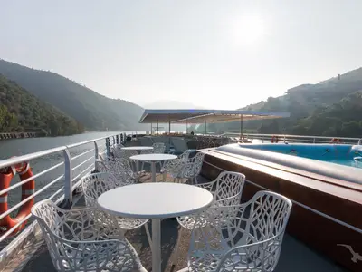 Das Sonnendeck der MS Magellan zeigt weiße Tische und Stühle, einen blauen Pool und eine malerische Flusslandschaft mit grünen Bergen unter hellem Himmel.