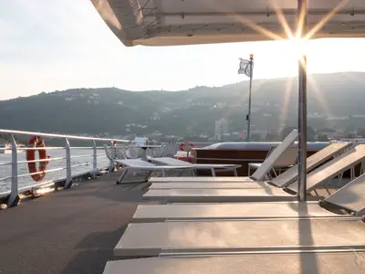 Das Sonnendeck der MS Magellan mit Liegestühlen und Tischen bei Sonnenuntergang. Im Hintergrund sind grüne Berge mit Bebauung und ein Fluss sichtbar.