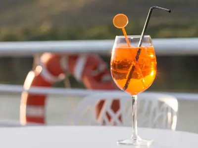 Nahaufnahme eines leuchtend orangefarbenen Cocktails mit Strohhalm und Rührer auf dem Sonnendeck der MS Magellan. Der Hintergrund ist unscharf und zeigt das Deck bei Sonnenschein.