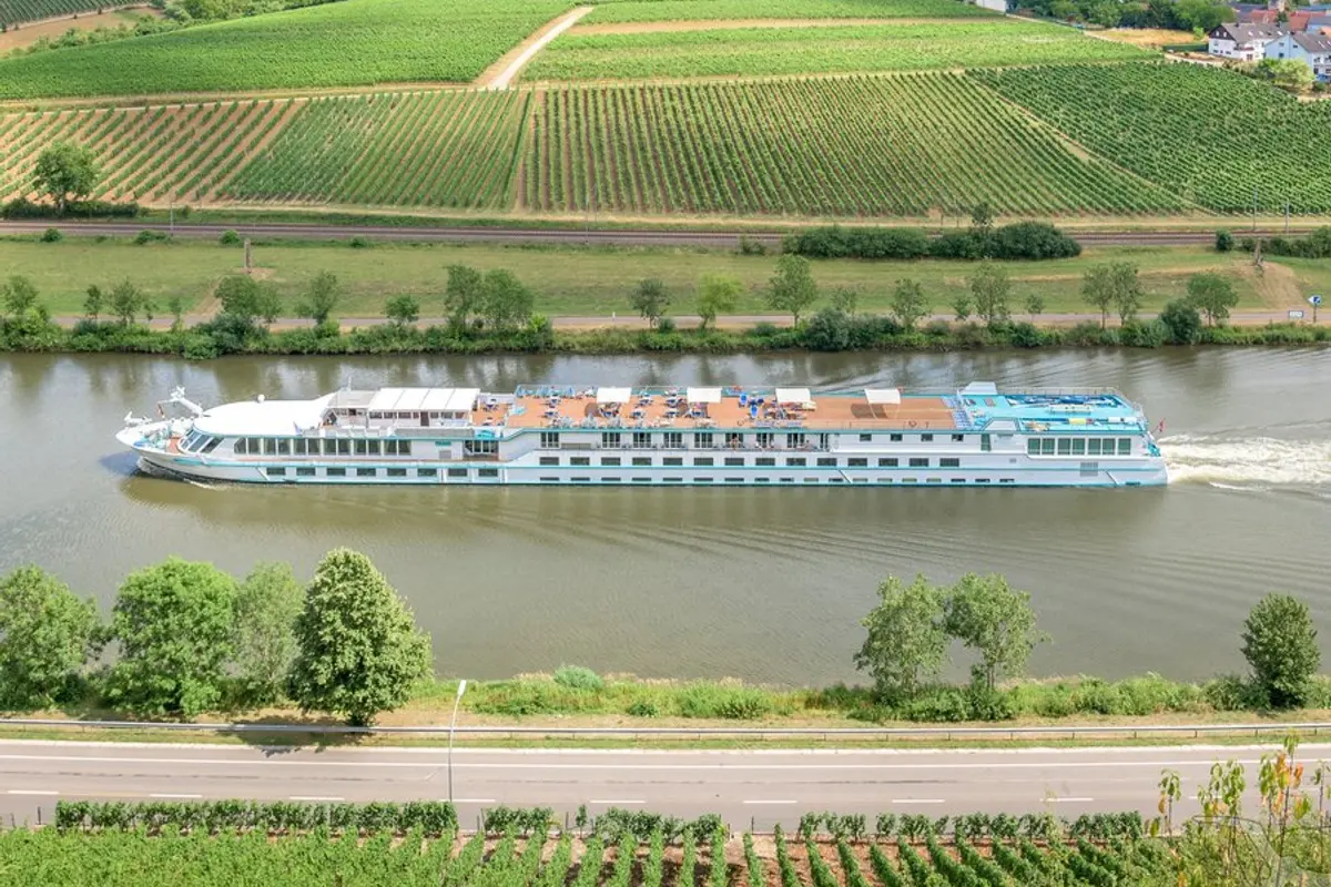 Die MS Rhein Prinzessin fährt auf einem Fluss, umgeben von grünen Weinbergen und Feldern. Das Sonnendeck zeigt viele Liegestühle.