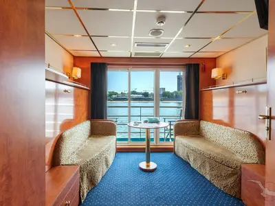 Balkonkabine der MS Rhein Prinzessin mit zwei Sofas und Tisch. Großes Fenster mit Blick auf den Fluss und die Stadt. Warme Holztöne und blauer Teppich prägen die gemütliche Einrichtung.