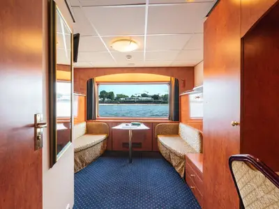 Helle Außenkabine an Bord der MS Rhein Prinzessin mit Sitzgelegenheiten. Ein Fenster bietet Flussblick auf eine grüne Uferlandschaft mit Bäumen und Gebäuden.