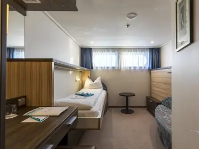 Blick in eine freundliche Außenkabine der MS Rhein Prinzessin. Ein Einzelbett, ein Schreibtisch mit Stuhl und zwei Fenster mit Vorhängen sind zu sehen.