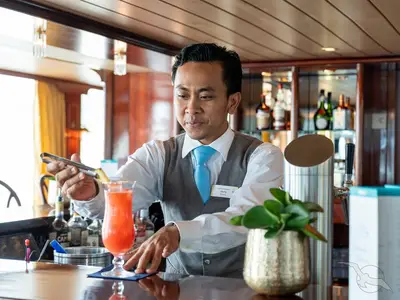 Ein Barmitarbeiter mit Namensschild "Agung" garniert an der Bar der MS Rhein Prinzessin einen roten Cocktail. Im Hintergrund sind Barregale mit Flaschen.