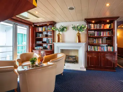 Helle Bibliothek an Bord der MS Rhein Prinzessin mit dunkelbraunen Bücherregalen, einem weißen Kamin und mehreren hellbraunen Ledersesseln. Ein großer Fensterausblick ist links sichtbar.