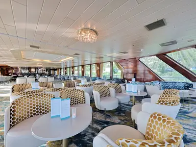 Helle Panorama-Lounge an Bord der MS Rhein Prinzessin. Elegante Sessel mit goldenen Mustern und Sofas an Tischen. Große Fensterfront mit Blick auf grüne Weinberge und Flusslandschaft. Ohne Personen.