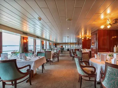 Elegantes Restaurant an Bord der MS Rhein Prinzessin mit weiß gedeckten Tischen und stilvollen Stühlen. Große Fenster bieten einen weiten Blick auf das Wasser. Warme Beleuchtung akzentuiert das klassische Ambiente.