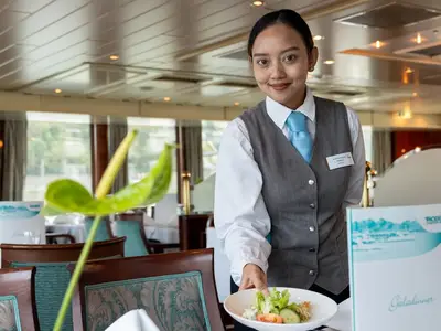 Eine lächelnde Crew-Mitarbeiterin der MS Rhein Prinzessin in grauer Weste und blauem Halstuch blickt in die Kamera. Im Hintergrund ist ein helles Restaurant mit großen Fenstern zu sehen.