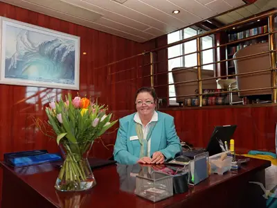 Lächelnde Crew an Rezeption der MS Rhein Prinzessin. Blumen auf dem Tisch. Im Hintergrund Bibliothek und Wellenbild an Holzwand.