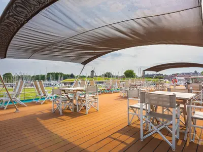 Das möblierte Sonnendeck der MS Rhein Prinzessin unter Sonnensegel. Im Hintergrund sind ein Hafen mit Segelbooten, eine Windmühle und Stadtgebäude zu sehen.