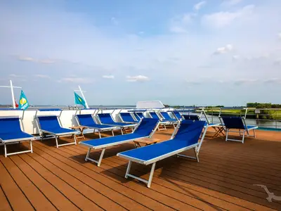 Sonnendeck der MS Rhein Prinzessin mit blauen Liegestühlen auf Holzboden. Flusslandschaft mit grünen Ufern unter blauem Himmel. Zwei Fahnen wehen an den Masten.