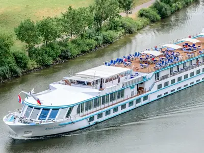 Vogelperspektive auf die MS Rhein Prinzessin, die auf einem grünen Fluss fährt. Viele Gäste entspannen auf dem Sonnendeck mit Liegestühlen und Tischen.