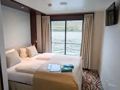 Gemütliche Kabine der MS Swiss Crystal mit zwei Betten und einem Fenster. Blick auf Fluss und Weinberge. Reiseunterlagen und kleine blaue Taschen auf dem Bett.