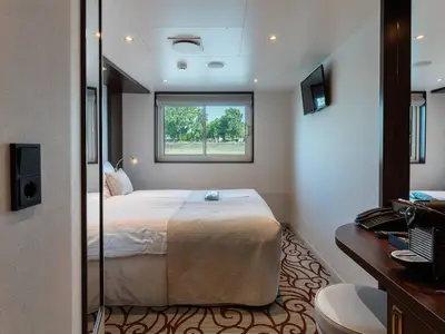 Helle Außenkabine an Bord der MS Swiss Crystal. Doppelbett, Fenster mit Blick auf Bäume und Wasser. Ein TV und Kaffeemaschine auf einem Schreibtisch sind sichtbar.