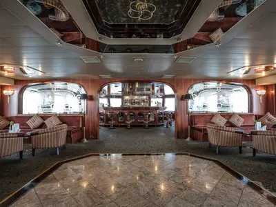 Blick in die elegante Bar-Lounge der MS Swiss Crystal. Eine zentrale Holzbar, umgeben von Sofas und Sesseln auf Teppich. Klassisches Design mit Marmorboden und Holzvertäfelung.
