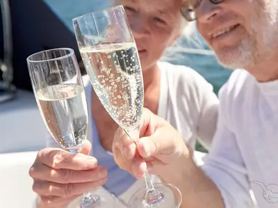 Zwei Personen stoßen auf der MS Swiss Crystal mit Sektgläsern an. Der prickelnde Champagner ist deutlich sichtbar. Im Hintergrund blaues Wasser. Ein festlicher Moment an Bord.